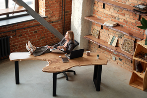 Above view of serious introspective mature woman in suit keeping legs on table while working with files in office