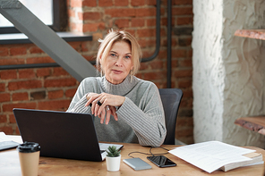 Portrait of content lady boss in gray sweater sitting at desk with laptop in modern loft office