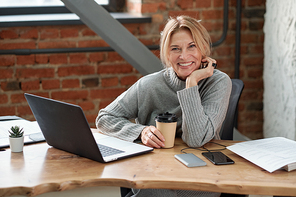 Portrait of excited business lady in gray sweater sitting at wooden table in office and drinking coffee
