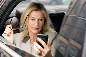 Blond serious mature woman reading message in smartphone while sitting on backseat of car or taxi cab by open window and hurrying for meeting