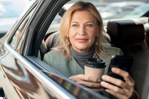 Mature woman having coffee and scrolling in smartphone while sitting on backseat of taxi cab and looking through open window