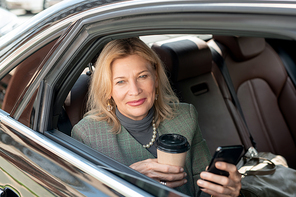 Mature woman with glass of coffee using phone while sitting on backseat of taxi cab and looking through open window on her way to work