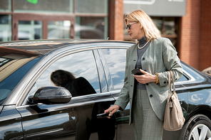 Elegant mature businesswoman or agent in sunglasses, grey suit and pearl necklace standing by black car and going to open door and get inside