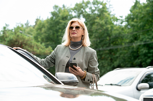 Elegant mature female business agent in sunglasses, suit and pearl necklace using smartphone while standing by car outdoors