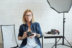 Portrait of female photographer in denim outfit sitting on stool in photo studio full of light