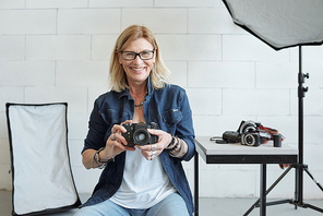 Portrait of cheerful mature photographer in eyeglasses sitting in photo shoot and using camera