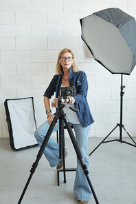 Portrait of cheerful mature photographer in eyeglasses sitting in photo shoot and using camera