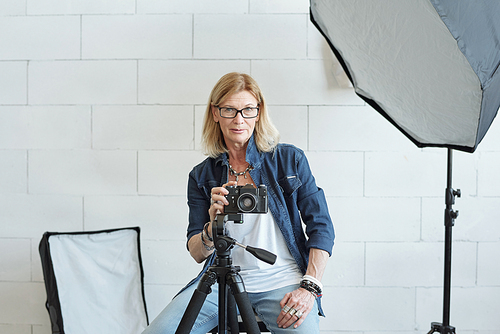 Portrait of serious photographer in eyeglasses adjusting camera on tripod while working in photo studio