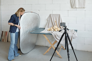 Mature photographer in denim outfit adjusting lighting reflector while preparing for photo shoot of food composition