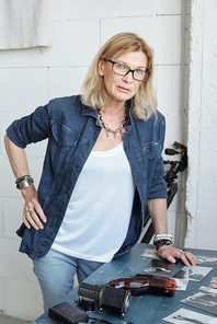 Portrait of confident mature photographer in denim jacket standing at table and choosing photos for album