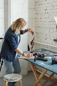 Blond-haired photographer in denim jacket standing at table with photographic equipment and choosing footage on film tape