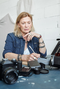 Blond-haired photographer in denim jacket standing at table with photographic equipment and choosing footage on film tape