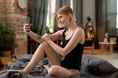 Happy young blond woman in black leotard making selfie or recording new video for blog on smartphone while sitting on bed after sleep