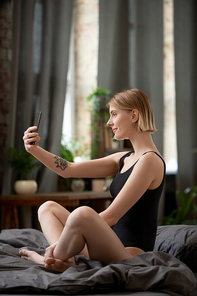 Happy young female in black leotard making selfie or recording new video for blog on smartphone in the morning while sitting on bed after sleep