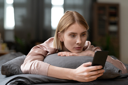Young blond female in beige silk robe or pajamas making selfie or scrolling through online news in the morning while relaxing on bed after sleep
