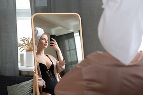 Young fresh woman in black lingerie and beige silk pajamas standing in front of large mirror in bedroom and making selfie after morning shower