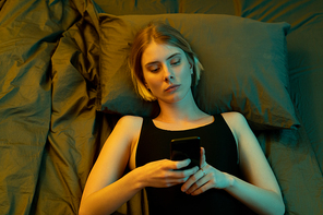 Young blond serious female in black leotard scrolling through online news in the morning or evening while lying on bed and keeping head on pillow