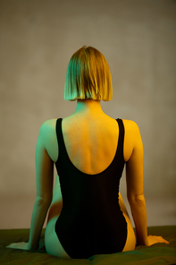 Rear view of young blond slim woman in black leotard sitting on bed in front of camera against grey wall in bedroom or studio