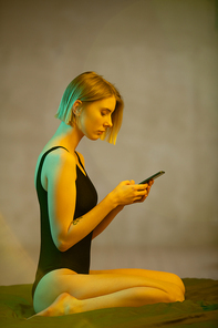 Side view of young blond serious female in black leotard scrolling through online news in the morning or evening while sitting on bed in isolation