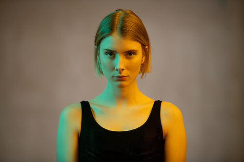 Young gorgeous blond female in black vest or leotard looking at you while standing in front of camera in isolation on background of grey wall