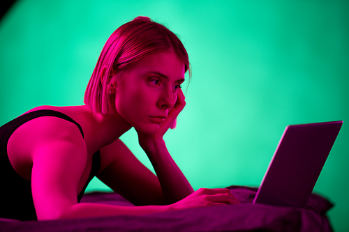 Blond serious girl in black underwear looking at laptop display while networking or watching online video on bed in the morning or evening