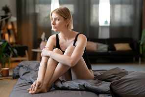 Young gorgeous blond female in black leotard keeping legs crossed while sitting on bed on Saturday morning and thinking of plans for the day