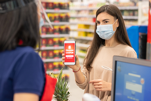 Modern woman wearing mask on face using digital discount coupon on her smartphone in supermarket