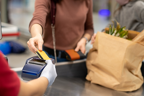 Hands of mature female buyer with smartphone over payment machine going to pay for food products in supermarket by cash register