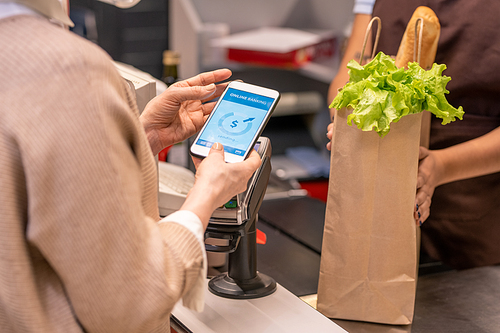 Hands of mature female buyer with smartphone over payment machine going to pay for food products in supermarket by cash register