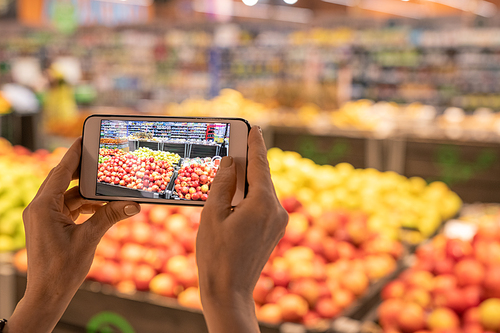 Hands of contemporary customer of supermarket holding smartphone and taking photo of display with fresh fruits and vegetables