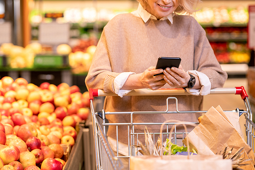 Mature female buyer in casual pullover scrolling through shopping list in smartphone while pushing cart with paperbags in supermarket