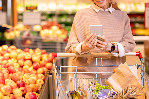 Mature female consumer in knitted beige sweater pushing cart with food products in supermarker and scrolling in smartphone