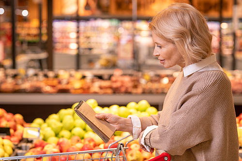 Pretty mature blond woman with notepad over cart reading shopping list while walking along display with fresh fruits in supermarket