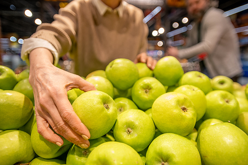 Hand of mature female consumer taking fresh granny smith apple while standing by heap of fruit during visit to supermarket