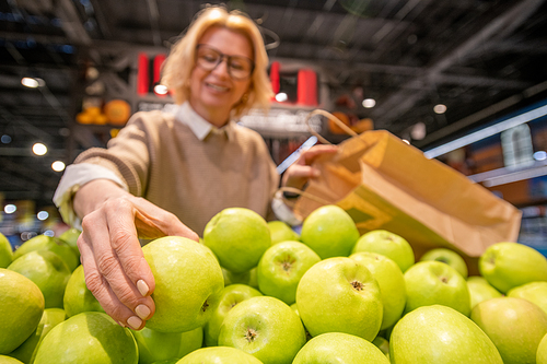 Blond mature female customer with paperbag choosing fresh green apples on fruit display while visiting supermarket and buying food