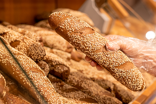 Gloved hand of mature female customer taking fresh bread with sesame sprinkles while buying food products in modern supermarket