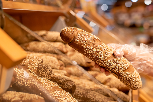 Gloved hand of mature female buyer holding fresh bread sprinkled with sesame seeds while standing by display in supermarket