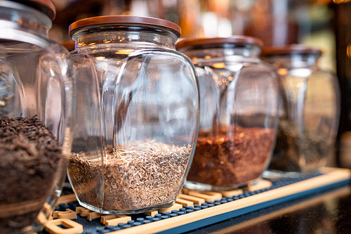 Row of big jars with fresh black, white, green and rooibos tea assortment standing on shelf in contemporary cafe or restaurant