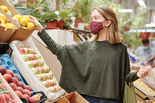 Attractive young woman in mask standing at counter and shopping for groceries during coronavirus