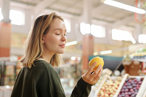 Content pretty girl standing at fresh market and looking at lemon while buying it