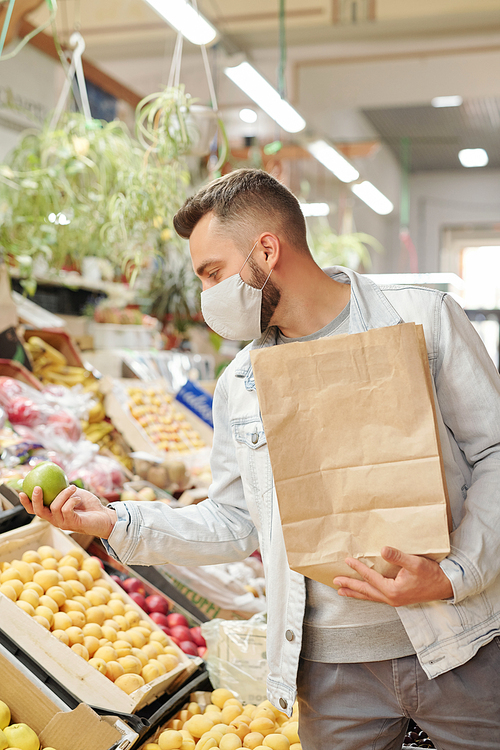 Young male customer in cloth mask standing at food counter and holding paper bag while buying apples at farmers market
