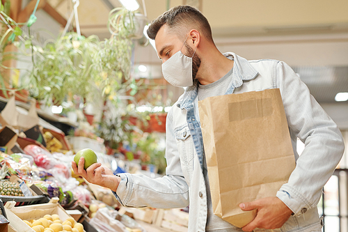 Young man in facial mask holding paper bag and choosing fruits while buying fresh groceries at organic market