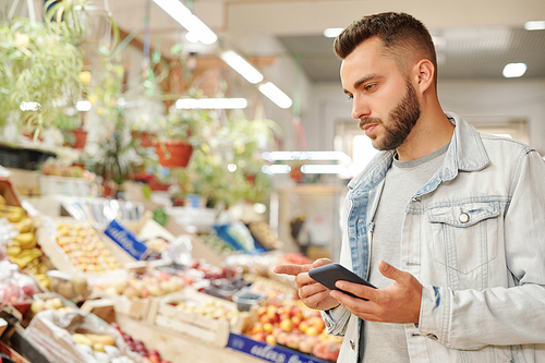 Serious young bearded man in jacket standing at food counter and checking groceries list via smartphone app