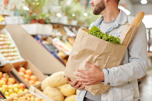 Close-up of bearded man in denim jacket carrying full paper bag of fresh products at food market