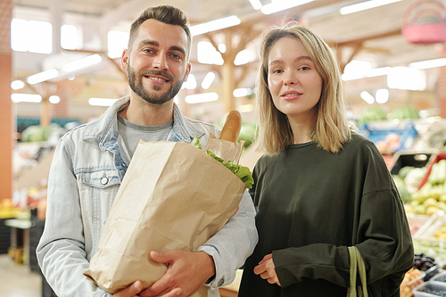 Portrait of positive young couple standing at farmers market and purchasing organic food together