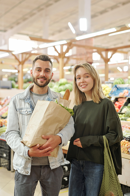 Portrait of smiling beautiful young couple in casual outfits standing with bags at modern farmers market
