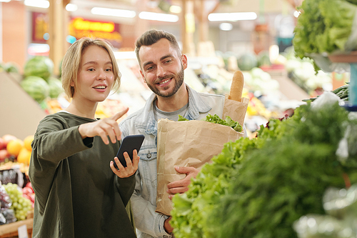 Positive young woman using smartphone app for purchasing and pointing at greens to boyfriend while they buying products at market