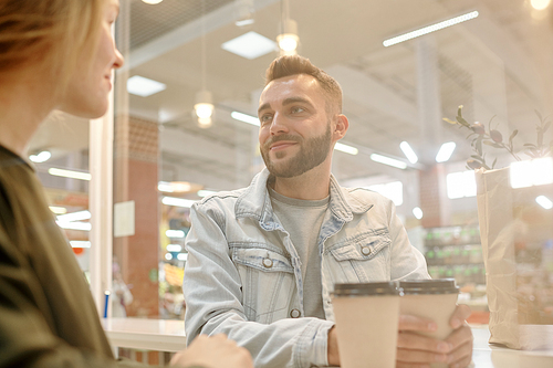 Handsome young bearded man in denim jacket sitting at counter and drinking coffee while dating with girl in fast food restaurant