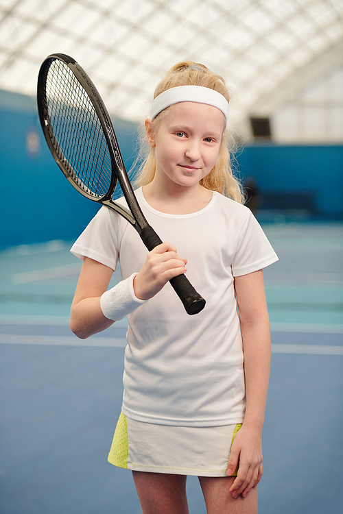 cute little girl with long blond hair holding tennis racket by right shoulder while standing in front of camera in modern stadium
