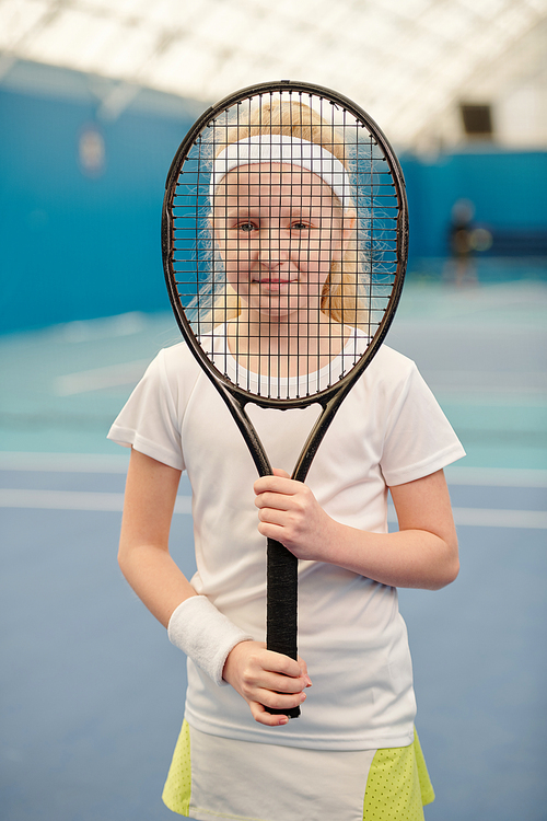 pretty blond girl in white activewear holding tennis racket in front of her face and looking at you through net in stadium